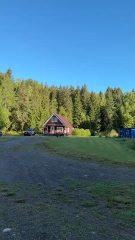 The video pans across a rural landscape with several red cabins nestled against a backdrop of dense green forest. A gravel driveway winds through the grassy area. In the foreground, the camera moves to the right, showing two cabins. One is a small, red house with white trim and windows. A picnic table is visible in front of it. Further to the right, another red cabin is partially visible. A black SUV is parked in the driveway of the first cabin. In the middle ground, a white motorhome is parked on the grass near a wooded area. Another black SUV is parked on the gravel road to the left of the motorhome. The sky is a clear, bright blue. The video captures a peaceful, natural setting with scattered dwellings.