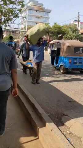 The video shows a man carrying a large green sack on his head, walking across a crosswalk in what appears to be an outdoor market area. He is wearing a blue shirt and brown pants, and sandals. Another man walks alongside him, pulling a small cart with coconuts on it. The scene is vibrant with people, tuk-tuks, and market stalls. The camera follows the man with the sack as he walks and crosses the street. The weather appears sunny and bright.