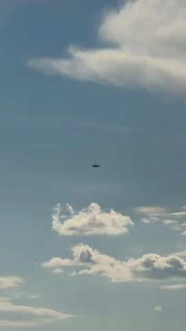 The video shows an airplane flying in a clear blue sky with scattered white clouds. The airplane is small and appears to be distant. The camera is pointed upwards, capturing the vastness of the sky. The lighting suggests it is daytime, likely afternoon due to the quality of the sunlight and the shadows on the clouds. There are no other objects or people visible in the frame. The airplane is the sole focus of the shot, moving slowly across the sky.