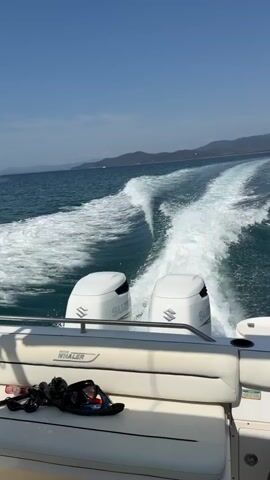 The video shows the back of a boat moving through the water, leaving a white wake. Two white outboard motors with "SUZUKI" branding are visible. The boat has white seating with a "WHALER" logo visible on it. There are some black items on the seat. The water is blue, and there are hills in the distance under a clear blue sky.