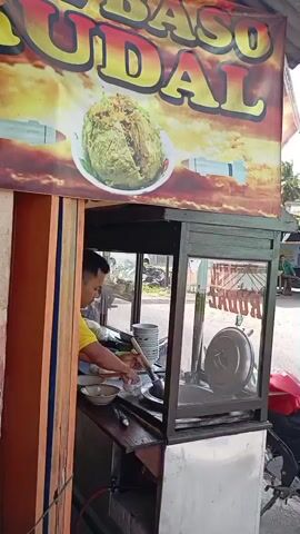 A person in a yellow shirt is preparing food at a street food stall. The stall has a banner with the text "BASO UDAI" and an image of a large ball of food. The person is holding a utensil over a bowl, possibly serving food. There are stacks of bowls to the left of the person. The stall is made of wood and metal, with glass panels. A red motorcycle is parked to the right of the stall. The background appears to be an outdoor setting with some trees and buildings visible.
