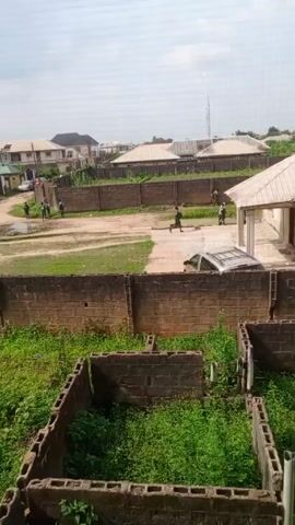 The video shows a daytime scene in a residential area, possibly a suburb or a developing neighborhood. The view is from an elevated position, perhaps from the upper floor of a building, overlooking a street and surrounding properties. Several one- and two-story houses with light-colored roofs and painted walls are visible in the background. A few vehicles, including a white pickup truck and a silver car, are parked or moving along the street. A group of individuals, appearing to be mostly young men, are gathered in the street, some walking and others standing in a cluster. They are dressed in casual clothing. The street is unpaved and appears muddy. The properties are surrounded by brick walls, some of which are covered with vegetation. In the foreground, there are some unfinished structures or foundations made of bricks, filled with overgrown grass and plants. The sky is overcast, suggesting a cloudy day.