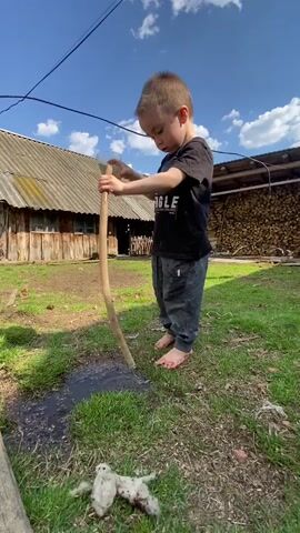 A young boy stands outdoors in a grassy area. He is barefoot, wearing grey sweatpants and a black t-shirt. He is pouring water from a clear plastic bottle into the top of a long, curved, wooden pipe. The water runs through the pipe and exits at the bottom, creating a puddle on the ground.  In the background is an old wooden building with a corrugated metal roof. To the right of the building is a stack of firewood. The sky is blue with scattered white clouds. A wire runs overhead. In the lower left corner of the frame, there is a white object on the grass. The boy focuses intently on pouring the water, and appears to be engaging in playful activity. Someone is talking in the background but is out of frame.