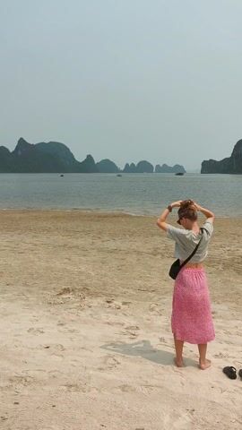 A woman stands on a sandy beach facing the sea, with islands in the background. She is wearing a light gray cropped t-shirt, a pink floral skirt, and a black shoulder bag. She is putting her hair up in a bun. Her sandals are on the sand beside her. The beach is wide and sandy, with some darker patches near the water. The sea is calm. The sky is overcast.