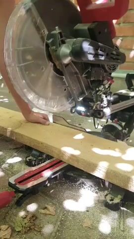 A person's hands operate a red and black miter saw. The saw is cutting a light-colored wooden plank. Sawdust is flying from the cut. The scene appears to be outdoors, with a brick wall in the background and dappled sunlight filtering through leaves overhead.