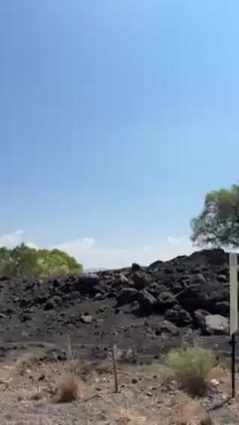The video captures a view from a moving vehicle, passing by a dark, rocky terrain with sparse vegetation under a clear, blue sky. As the vehicle progresses, trees come into view, partially obscuring a distant body of water and low hills. The ground close to the road appears dry and somewhat barren.