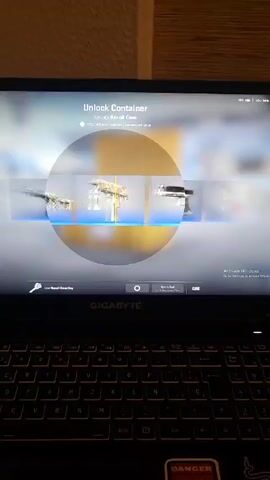 The video shows a laptop screen displaying the Counter-Strike: Global Offensive (CS:GO) case opening animation. The user is unlocking a Recoil Case. After the animation completes, the revealed item is an SG 553 Dragon Tech. The laptop is a GIGABYTE model with a sticker at the bottom right corner of the keyboard area that says "DANGER". The laptop is on a surface with a wooden-looking background. The Windows operating system on the laptop needs to be activated.
