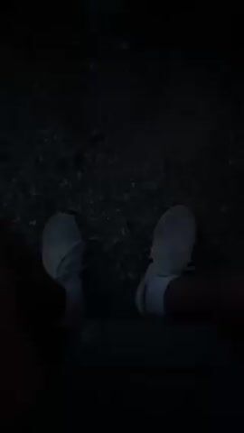 Here is a detailed description of the video:

The video starts with a low-angle shot of someone's feet in white shoes, situated near what appears to be a low stone wall or ledge. The lighting is very dim. The camera then tilts upward to reveal a stone fire pit filled with a lively fire. The backdrop transitions from near darkness to the early stages of twilight, with the sky turning a deep blue. Trees and the faint outline of a house with illuminated windows can be seen in the background.

The video continues to pan out, showing three people sitting around the fire pit. One person stands and adjusts their clothing. The group appears to be enjoying the fire and the outdoor setting. The video concludes with the camera focusing back on the person's legs who recorded the video.