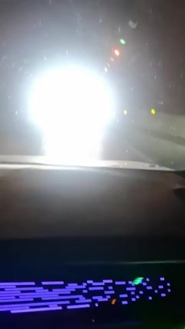 A car exits a tunnel and drives along a mountain road. The mountains are rocky and green, with some snow on their sides. There are two trucks and a car ahead on the road.  The car is filming from the driver's perspective.  The sky is blue with some clouds.