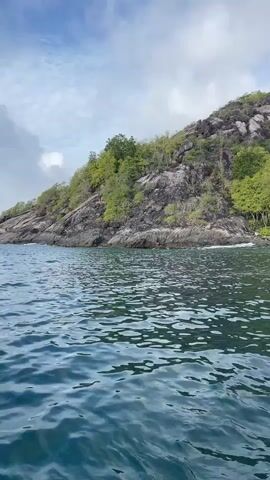 The video shows a body of water in the foreground, with gentle ripples. In the background, a rocky, tree-covered hill rises steeply from the water's edge. The sky above is partly cloudy, with patches of blue visible. The scene appears to be a coastal or island landscape. There are no people or specific objects clearly visible, other than the natural elements of water, rock, and vegetation. The perspective suggests the camera is on the water, possibly from a boat, looking towards the shore.