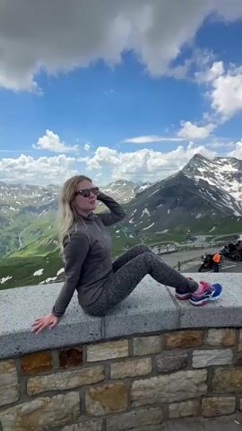 A woman with blonde hair and sunglasses is sitting on a stone wall in a mountainous landscape. She is wearing a grey zip-up jacket and patterned leggings. Her legs are crossed and she is looking up at the sky. Behind her, there are green hills, snow-capped mountains, and a blue sky with white clouds. There are also motorcycles parked to the right of the frame. The video appears to be a short, static shot with some subtle camera movement.