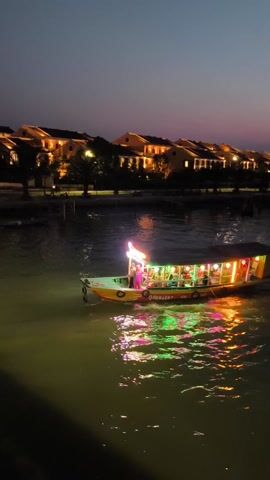 At night, a colorful boat cruises on a river past buildings. The boat is lit with red, yellow, and green lights, and it has a canopy over the seating area. There appear to be people sitting on board. The boat’s reflection is mirrored in the water. The boat’s name, ONANH 297, is emblazoned on its side. The buildings on the shore are brightly lit. The sky is dark, and the river water appears greenish. The video is shot from a slightly elevated position.