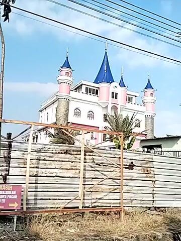 The video captures a moving street view in Sidoharjo, Indonesia, under a bright, sunny sky with clear blue conditions and scattered clouds.

Initially, the camera focuses on a distinctive building resembling a fairytale castle. It features pink and white walls, blue pointed roofs (turrets), and numerous windows. Prominent power lines and utility poles crisscross the sky in the foreground, with a small green bush visible.

The scene then transitions to an architectural complex featuring predominantly white and yellow buildings. These structures are adorned with multiple green domes, suggesting a mosque or Islamic institution. The camera approaches a main entrance where a large sign is clearly visible. The sign reads "KAMPUS AZHAR" and "AL AZHAR" in large lettering, with smaller text underneath that appears to say "SIDOARJO - MOJOKERTO." An arrow points left, accompanied by the word "MASUK" (Entry).

The entrance area includes landscaped greenery with trees, and a red flag flies on a flagpole in the distance. A road with black and white striped curbs runs in front of the campus. Power lines and utility poles continue to be a visible element along the street. No individuals are seen in the video.