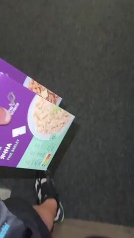 A person stands on a gray carpet, holding two purple and green boxes of "Krupa Yachna Fine Barley" in their left hand. They are wearing black shorts and black and white sneakers with green accents. In the background, against a white door frame, are a pair of clear jelly sandals. The person pivots slightly, keeping the boxes in view.