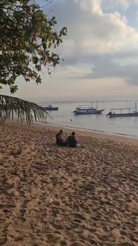 The video shows a wide shot of a sandy beach with two men sitting on the sand facing the ocean. They are both wearing black tank tops and shorts. In the background, there are several boats floating on the water, and a person can be seen swimming in the ocean. The sky is partly cloudy, and the lighting suggests it is either early morning or late afternoon. There are some palm fronds hanging from the top left of the frame, indicating a tropical location.