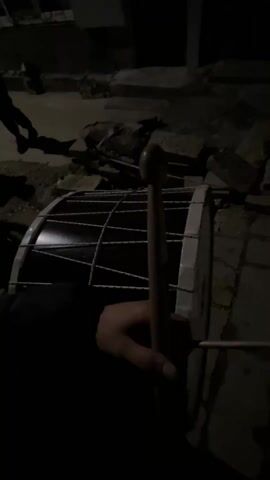 At night, a person holds a stick next to a large drum, preparing to play. The surroundings are dark, with cobblestones underfoot and hints of foliage and a few other people in the background. A parked car is partially visible. The street is dimly lit by a distant street lamp.