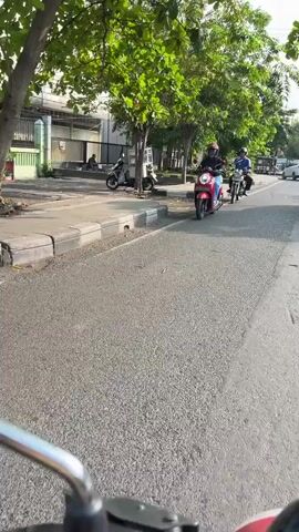 The video is filmed from the perspective of someone riding a red scooter. In the foreground, the handlebars of the scooter are visible. The road ahead is paved asphalt. To the left of the road, there is a sidewalk with a curb. Behind the sidewalk, there are several parked motorcycles and a building with a green fence. Further down the road, a person is riding a red scooter, followed by two other people on motorcycles. Trees with green foliage line the left side of the road. The scene appears to be a sunny day in an urban environment.