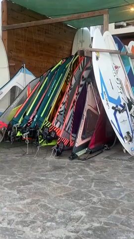 The video shows a rack of windsurfing boards and sails leaning against a wall. The boards are of various colors and designs, and the sails are also colorful. The camera pans slowly across the rack, showing the equipment in detail. The ground is paved with stone tiles, and there is a wooden structure overhead.