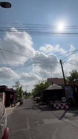 The video pans across a street with houses on either side. The sky is bright blue with fluffy white clouds, and the sun is shining. There are many overhead wires crisscrossing the street. On the left side of the street, there is a person on a motorcycle. On the right side, there is a small covered structure with a red and white checkered barrier in front of it. Further down the street, there is another person pushing a cart loaded with goods. Trees and lush greenery are visible throughout the scene.