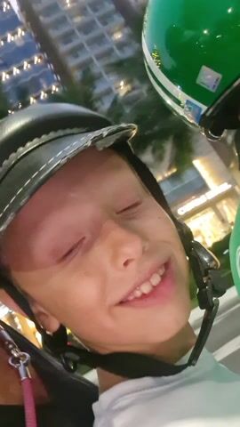 A woman and a young boy are on a motorbike. The woman sticks out her tongue and smiles. The boy smiles. They are both wearing black helmets. The woman has dark hair and is wearing a pink strap around her neck. The boy has light hair and is wearing a white shirt. A green helmet is visible in the background. Buildings and palm trees are also visible in the background.