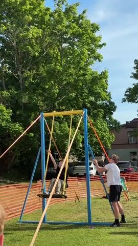 A person is performing an aerial stunt on a swing-like apparatus. They are upside down, holding onto two vertical yellow poles with their hands. The poles are suspended from a horizontal yellow bar. The apparatus is supported by a blue metal frame. Several orange and yellow straps extend from the yellow bar down to the ground, and a person in a white t-shirt and black shorts stands below, holding one of the straps, likely to assist or control the movement. The stunt takes place outdoors on a grassy area, with trees and a building visible in the background. The sky is blue with some clouds. A portion of another person's head and shoulder is visible in the lower left corner of the frame. The person performing the stunt is wearing a black t-shirt and light-colored pants.