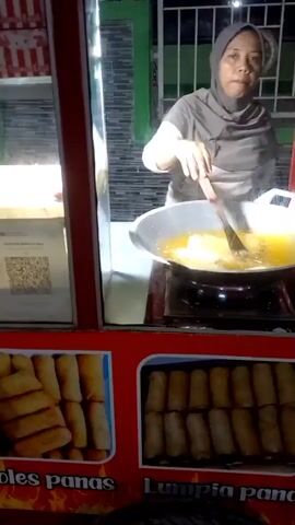 A woman wearing a grey headscarf and a light-colored shirt is standing behind a food stall and frying spring rolls in a large wok filled with oil. She is holding a wooden spatula and appears to be turning the spring rolls in the hot oil. The stall has a glass front, and the woman is visible through it. Below the cooking area, there are images of fried spring rolls displayed on the stall. The text on the stall, partially visible, includes "oles panas" and "Lumpia Panas," indicating hot spring rolls. There is also a QR code displayed on the glass, likely for payment or information. The background is dimly lit, suggesting it might be evening or nighttime.