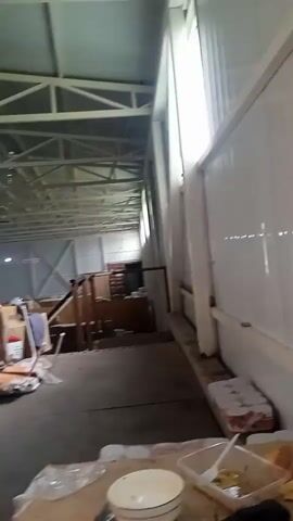 The video shows an interior scene, likely a warehouse or workshop, with the camera panning from right to left. The right side of the frame is dominated by a white, corrugated metal wall, possibly an exterior wall or internal partition. A window with a white frame is visible in the upper right section. In the foreground, on a brown cardboard surface, are remnants of a meal. This includes a white plate, a clear plastic container with some food and a fork, and a couple of hamburger buns. To the left of the meal, a stack of white items, possibly paper towels or toilet paper rolls, is visible.

The camera then pans left, revealing a doorway with a white door and a glass panel, situated within a wall constructed of oriented strand board (OSB). Further into the space, the view opens up to a long, dimly lit corridor. The ceiling appears to be a metal truss structure, with exposed beams and lighting fixtures. On the left side of the corridor, there are various objects, including stacked cardboard boxes, wooden shelves, and what looks like furniture or equipment covered with tarps. The overall impression is one of a functional, possibly storage or work area, with evidence of recent activity.