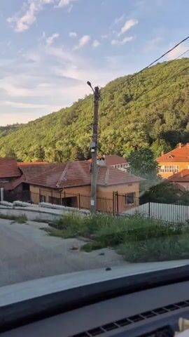 The video shows a view from inside a vehicle, looking out at a rural landscape. The vehicle is moving forward on a paved road that slopes slightly uphill. On the right side of the road, there is a pale orange house with a red-tiled roof and a black metal fence. A utility pole with wires is visible in front of the house. Behind the house, a lush, green, tree-covered hill rises steeply. On the left side of the road, there are other houses with red-tiled roofs and fences, and more vegetation. The sky is blue with scattered white clouds, and the lighting suggests it is daytime, possibly late afternoon or early evening due to the warm tones. The overall scene depicts a quiet, residential area nestled against a natural, hilly backdrop.