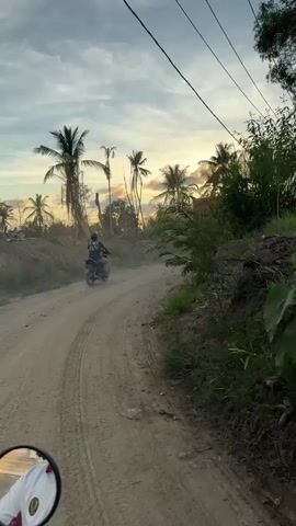 The video shows a first-person perspective from a moving motorcycle on a dusty dirt road. The sun is setting, casting a warm glow through palm trees and a hazy sky. A person on a motorcycle is ahead on the road, kicking up dust. Further ahead, a large orange dump truck is also moving forward, creating a significant dust cloud. The road is lined with vegetation and some debris. Power lines are visible overhead.