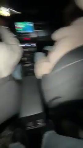 The video shows three women in the back seat of a car. The camera is held by one of them, pointing up at the faces of the group. The women are all wearing winter clothing, including jackets and hats. The woman holding the camera is in the middle, wearing a black beanie and a black jacket. The woman to her left has blonde hair and is wearing a brown fur-lined jacket. The woman to the right has long curly brown hair and is wearing a green scarf and a black jacket. They appear to be singing along to music playing in the car. The interior of the car is visible.