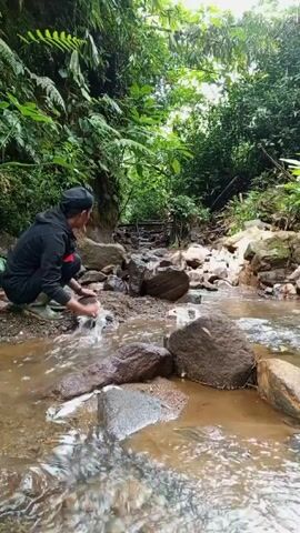 A man in a black outfit and boots crouches by a shallow, rocky stream, then stands up and walks further into the scene, towards a small set of wooden steps. The stream is surrounded by dense green vegetation.
