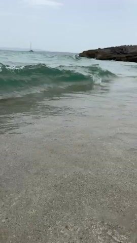 The video shows a close-up view of gentle waves breaking on a sandy beach. The water is a translucent blue-green, and small bubbles form as the waves recede. In the background, a sailboat with a white sail is visible on the horizon, and a rocky outcrop juts into the sea on the right side of the frame. The sky is overcast, suggesting a cloudy day. The camera is positioned at a low angle, close to the water's edge, emphasizing the movement and texture of the waves.