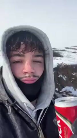 A person in a snowy landscape drinks from a red and silver can. The person wears a grey hoodie, a black jacket, and black gloves. They have curly hair and a bit of facial hair. The background shows a snowy hill with a road, a car parked on the side, and a rainbow in the sky. The weather appears cold and bright. The person takes a sip from the can and then looks around at the scenery.
