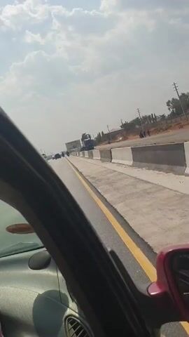 The video shows a view from inside a moving car, likely on a newly constructed or reconstructed road. The road has concrete barriers along the sides and a yellow lane marking. In the distance, other vehicles, including large trucks, can be seen moving along the road. An advertising billboard is visible on the side of the road. The sky is partly cloudy. The video appears to be taken during the day, with ample sunlight.