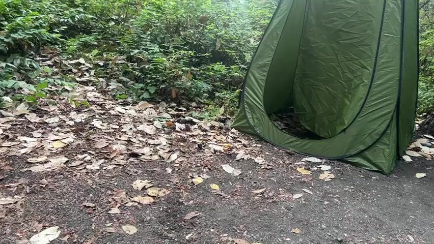 The video shows a portable green changing tent set up outdoors in a wooded area. The ground is covered with fallen leaves and dirt. The tent is open, revealing the dark interior. The background is filled with lush green foliage and trees. The camera pans slightly to the right, showing a large tree trunk next to the tent. A small bird is visible on the ground to the right of the tent. The lighting suggests an overcast day.