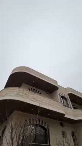 The camera looks up at a modern, multi-story building with rounded balconies and stone facing.  The sky is overcast.  Trees with bare branches are in the foreground. In the distance, other buildings are visible, as is a small hill.  The camera pans slightly to the left.