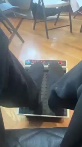 The video shows a person's feet in black socks on a foot massager. The massager is gray and white with a ribbed black surface. In the background, there is a person sitting in a massage chair near a window. The room has hardwood floors and several folding chairs. There is a glimpse of greenery visible through the window. The overall setting appears to be indoors, possibly a living room.