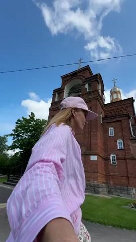 A first-person perspective video shows a young woman with blonde hair wearing a pink baseball cap and a pink and white striped button-down shirt. She is looking towards her right and upwards. Behind her is a large, red brick building with a bell tower, topped with crosses. The building appears to be a church. The sky is blue with some white clouds. The woman is standing on a paved surface, and there is green grass and some bushes to her right.