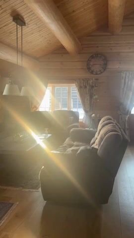 The video shows the interior of a wooden cabin or house. The walls and ceiling are constructed from logs. Sunlight streams in through a window on the far wall, creating a bright glare. There is a large, brown, plush sofa in the foreground and another armchair partially visible to its left. A coffee table with items on it is in front of the sofa. A large clock hangs on the wall above the sofa. Curtains frame the window behind the sofa. There is a rug on the floor near the window.
