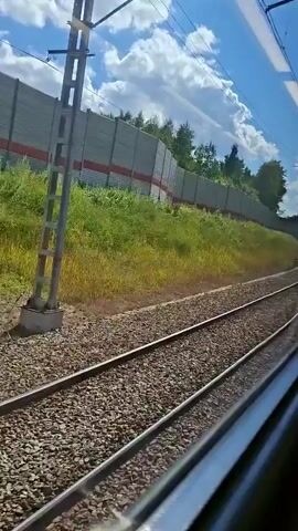 The video shows a view from a moving train. The camera is looking out of the side window, capturing the landscape rushing by. In the foreground, there are train tracks covered with gravel. To the left of the tracks, there is a grassy embankment topped with a tall sound barrier fence. The fence is made of grey panels with a red stripe near the bottom and appears to have some graffiti on it. Above the fence, there are trees and lush green foliage. Overhead, power lines with a supporting pole are visible against a bright blue sky dotted with white clouds. The lighting suggests it is daytime, possibly late morning or early afternoon, with the sun casting shadows and illuminating the scene. The train is clearly in motion, indicated by the blurred effect on the passing scenery. There are no people visible in the frame.