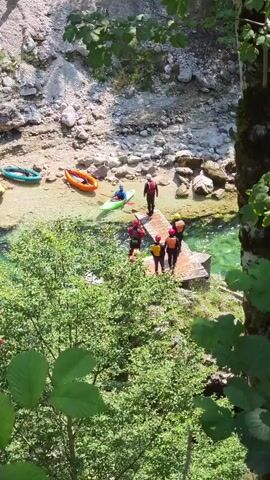 A group of people in kayaks and wetsuits are gathered on a wooden dock next to a clear, flowing river. Several kayaks are lined up on the sandy bank of the river. The area is surrounded by lush green trees and rocky terrain. The scene appears to be an outdoor recreational activity.