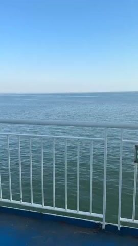 This media file shows a view from a ferry on a clear, calm day. The foreground features a white railing and a blue deck, suggesting the perspective of someone standing on the ferry's deck. Beyond the railing, the vast, blue expanse of the sea stretches to the horizon. The water is a deep, tranquil blue with gentle ripples, indicating minimal waves.

In the distance, a large cargo ship is visible, appearing as a small silhouette against the horizon. The ship is red and white, with a long hull. The sky above is a clear, pale blue, fading to a lighter hue near the horizon, which suggests it is either early morning or late afternoon.

The overall scene evokes a sense of peace and tranquility, with the open sea and the distant ship creating a feeling of solitude and journey. There are no people visible in the frame. The setting is clearly on a body of water, likely a sea or a large bay, given the presence of a cargo ship. No specific location cues are evident beyond this. A small green sign with white text, possibly a number, is partially visible on the white railing on the right side of the frame.