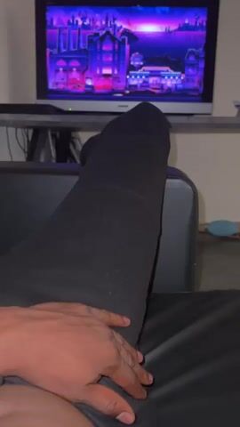 A first-person perspective video shows a person's legs and hand on a black couch, with a television screen in the background displaying a colorful animated scene. The person is wearing black pants and black socks. The television appears to be a flat-screen monitor on a stand, and the background is dimly lit. The video is brief, with no discernible action or interaction.