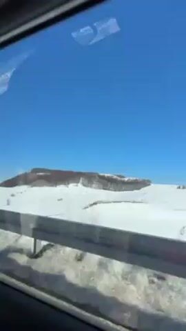 The video shows a view from inside a moving vehicle, likely a car, driving along a road next to a snowy field. The guardrail is visible in the foreground. Beyond the guardrail, the landscape is covered in snow under a clear blue sky. In the distance, there is a small hill or raised area with a few trees or bushes. The overall impression is one of a winter scene captured while traveling by car.