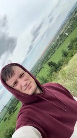 A person wearing a maroon hoodie with white sleeves poses for a selfie outdoors. The person's face is visible, and they are looking at the camera with a slightly strained expression. The background shows a landscape with rolling green hills, fields, and a cloudy sky. The camera angle is tilted.