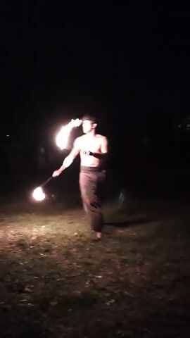 A person is performing with fire in a dark, grassy outdoor area. The person is holding two flaming objects, one in each hand, and is moving them in a circular motion. The flames are bright and illuminate the immediate surroundings. In one shot, the person exhales a large plume of fire. The person is wearing loose-fitting pants. In the background, there are trees and possibly other people sitting. The scene suggests a fire performance or show.