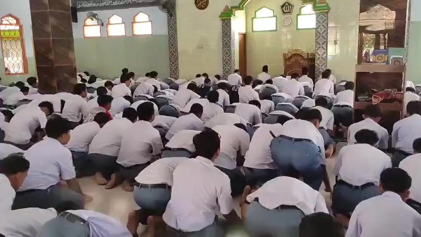 The video shows a large group of young males, most likely students, dressed in white shirts and dark pants or trousers, sitting on the floor of a mosque. They are facing towards the front of the mosque where a mihrab, a niche indicating the direction of prayer, is visible. Some individuals are standing, possibly instructors or religious leaders, moving among the seated students. The environment is an indoor space with tiled floors and walls adorned with religious calligraphy and decorative elements. Natural light enters through windows. The overall atmosphere appears to be one of prayer, religious observance, or instruction.