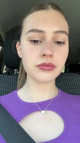 Here is a description of the video:
The video features a young woman with fair skin and light brown hair, pulled back into a ponytail. She is wearing a light purple ribbed shirt with a keyhole cutout at the chest and a silver necklace with a heart-shaped pendant. She has subtle makeup on, including lip gloss. She is wearing a seatbelt. The background shows the interior of a car with gray and black patterned seats. There is music playing in the background. She pulls her hair back behind her ear.