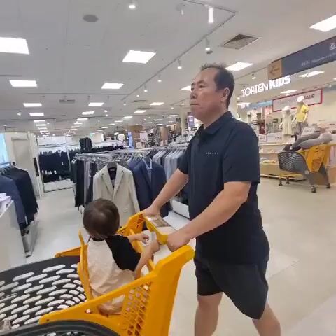 A man pushes a yellow shopping cart with a child sitting in the child seat. The man is wearing a black polo shirt and black shorts. The child is wearing a black and white shirt. They are in a clothing store. There are racks of clothes and mannequins in the background. The floor is tiled. The lighting is bright, likely from overhead fluorescent lights. The camera is mounted low, possibly on the shopping cart itself, giving a perspective from within the cart or just behind it.