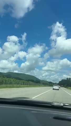 This is a first-person perspective video taken from inside a car driving on a highway. The camera is pointed forward, showing the road ahead and the sky above. The sky is a bright blue with scattered white cumulus clouds. In the distance, there are green hills or mountains. Along the side of the road, there are trees and what appears to be a vineyard or cultivated field. There are a few other cars visible on the highway, including a white car in front of the vehicle the video is being filmed from. The road has white lane markings and appears to be in good condition. The overall impression is of a sunny day, possibly in a rural or semi-rural area.